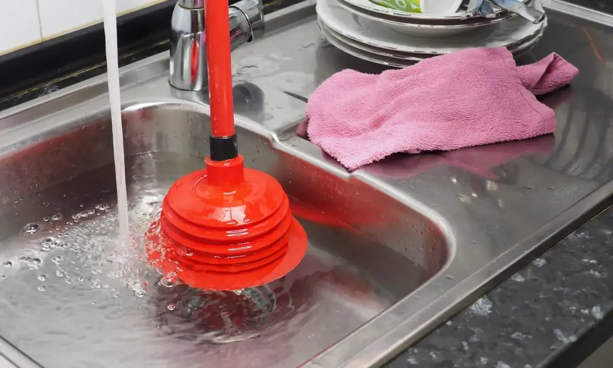Un desatascador casero rojo en un fregadero de acero inoxidable con agua corriendo. Hay platos sucios apilados y una toalla rosa.