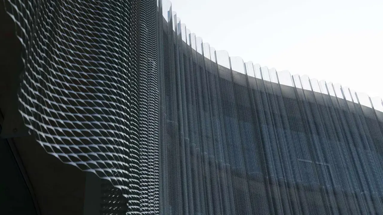 Detalle de una fachada ventilada con malla metálica expandida que crea un patrón ondulado y vertical, reflejando la luz.