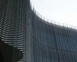 Detalle de una fachada ventilada con malla metálica expandida que crea un patrón ondulado y vertical, reflejando la luz.