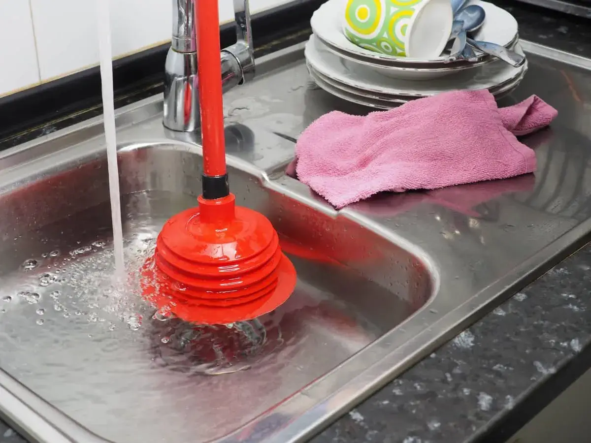 Un desatascador casero rojo en un fregadero de acero inoxidable con agua corriendo. Hay platos sucios apilados y una toalla rosa.