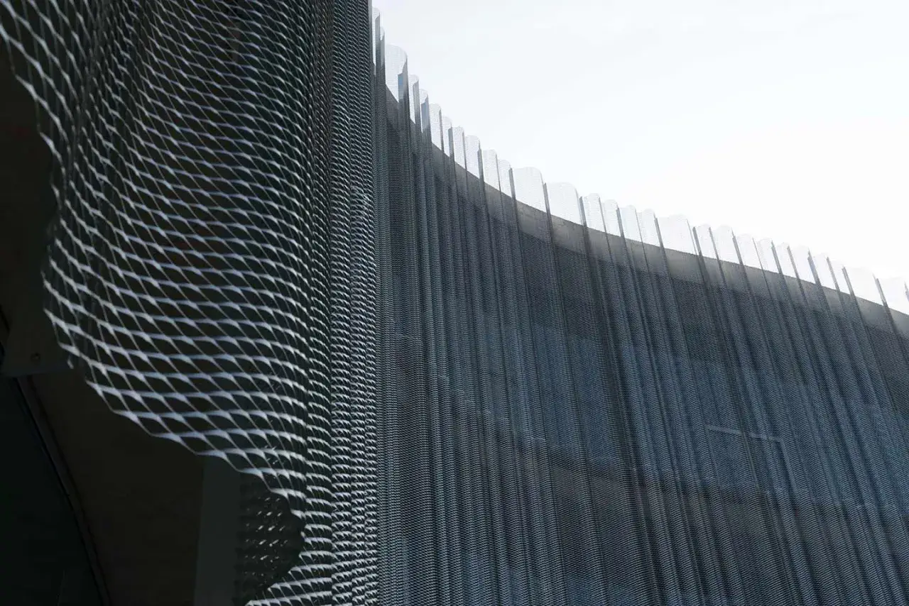 Detalle de una fachada ventilada con malla metálica expandida que crea un patrón ondulado y vertical, reflejando la luz.