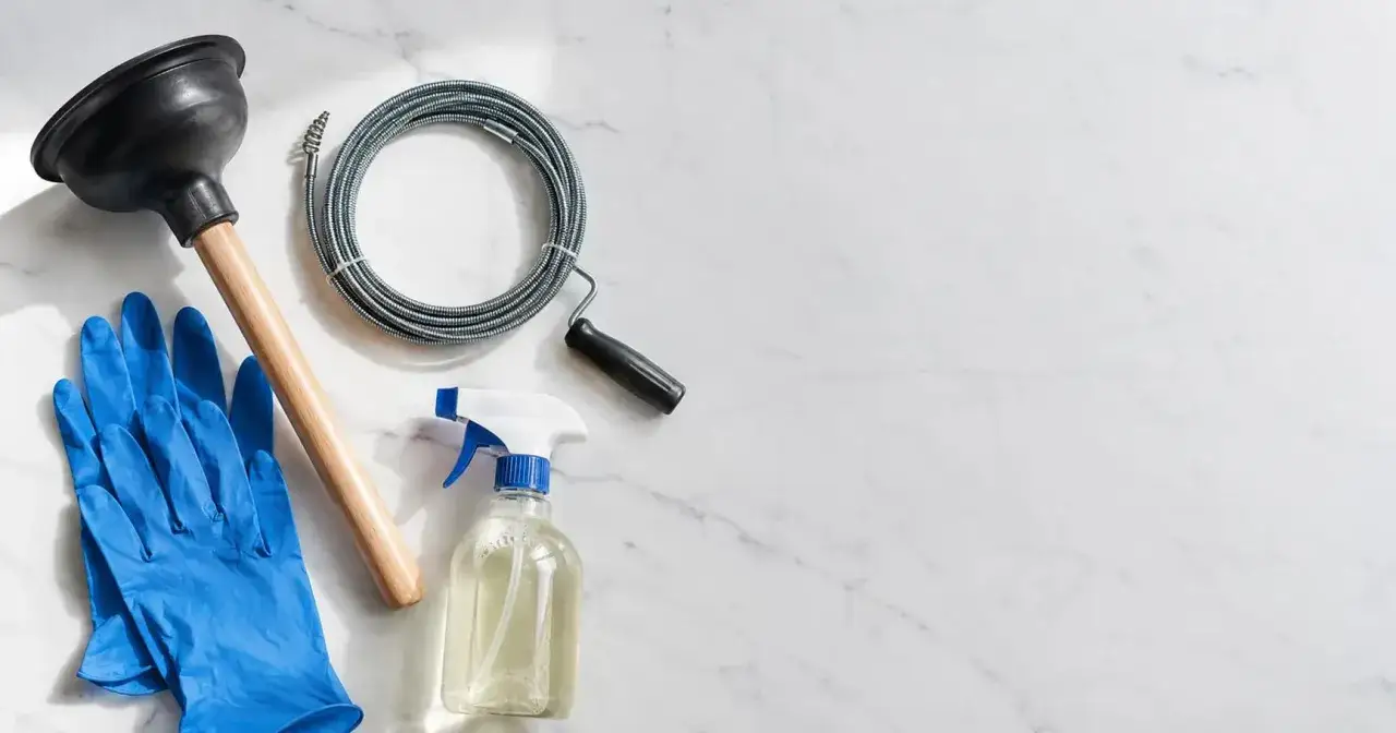 Un desatascador casero, guantes azules, un cable de desatasco y un spray limpiador sobre una superficie de mármol.