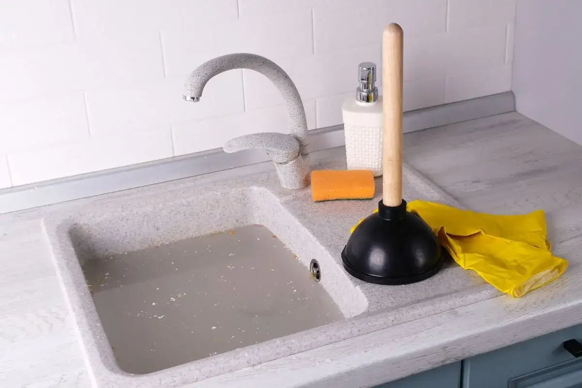Un fregadero atascado con agua sucia y restos. Un desatascador, guantes amarillos y una esponja listos para desatascar fregadero.