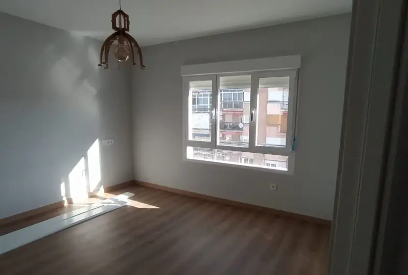Habitación con suelo de madera, paredes grises y ventana. El sol ilumina la estancia, sugiriendo un buen **precio medio reforma integral**.
