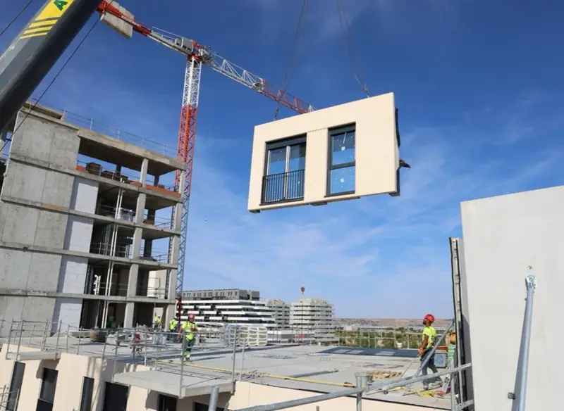 Grúa elevando un módulo prefabricado para la construcción industrializada de un edificio. Obreros supervisan la operación.
